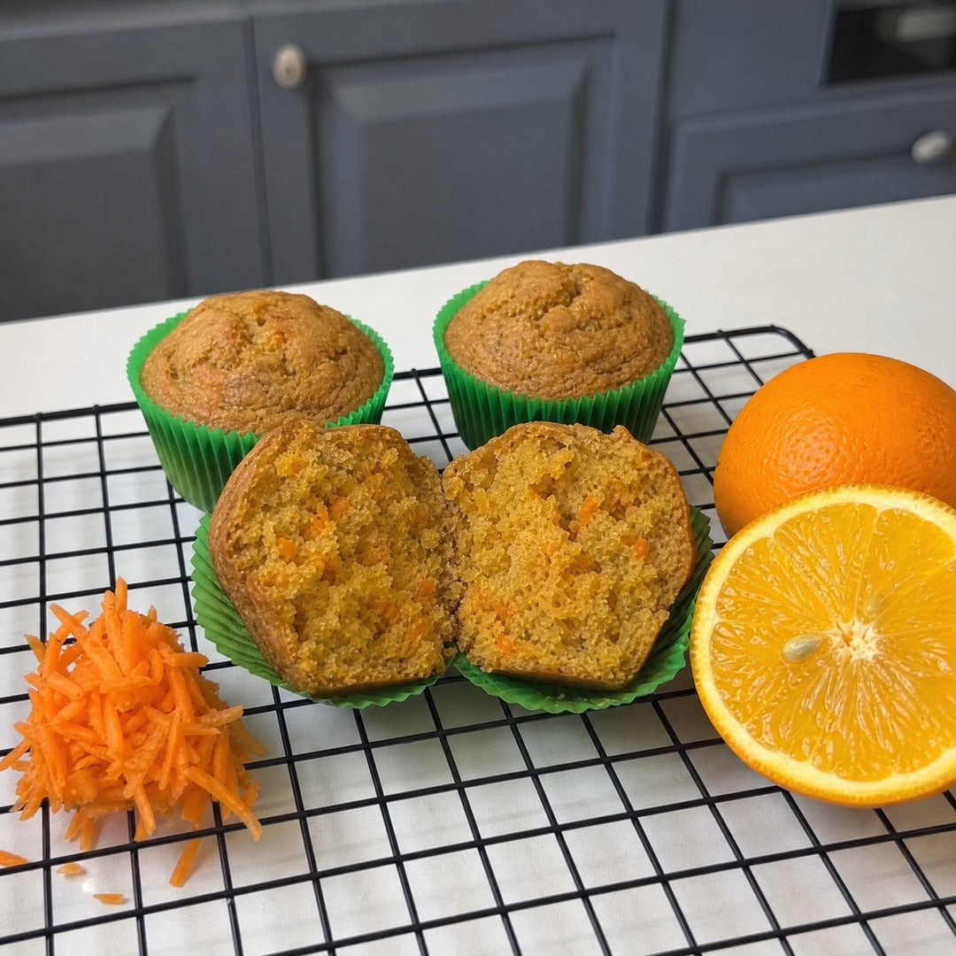 Muffins de cenoura em forminhas verdes sobre grade de metal, um partido ao meio mostrando o miolo alaranjado, ao lado de laranja e cenoura ralada