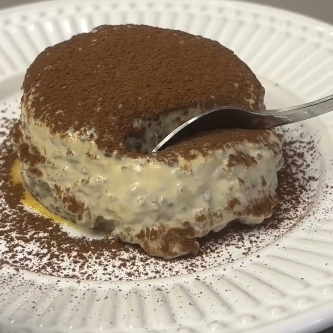 Tiramisú de chia coberto de cacau em pó num prato branco, com a colher rompendo a camada escura e mostrando o creme claro por baixo